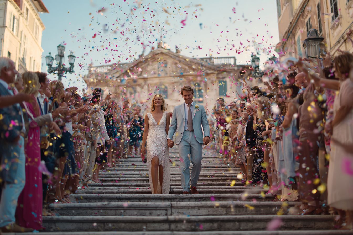 couple de mariés sortant d’une mairie française en pierre claire, escalier monumental, invités alignés des deux côtés lançant confettis multicolores et rubans
