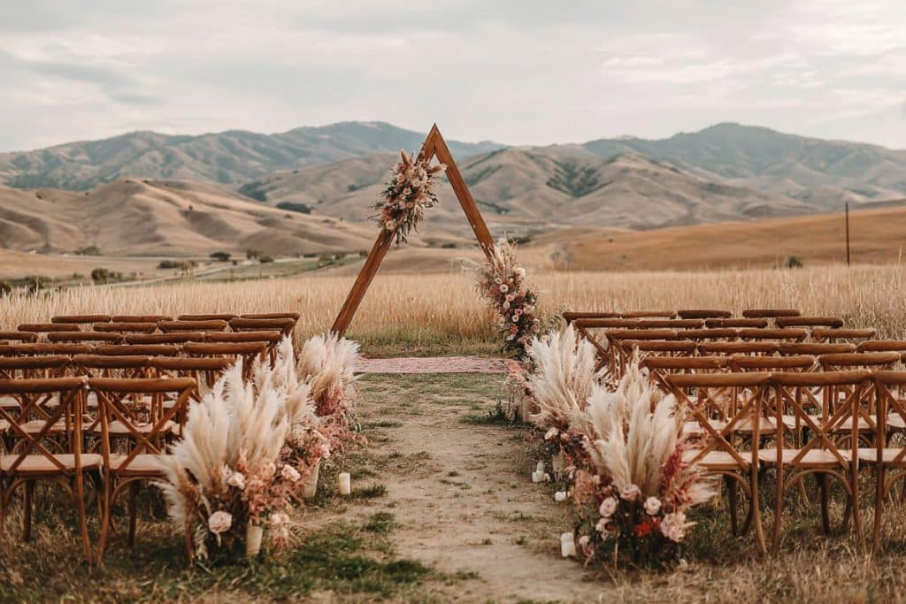 Installation de cérémonie laïque pour un mariage dans un champ, avec une allée de chaises en bois dépareillées et une arche bohème décorée de fleurs séchées
