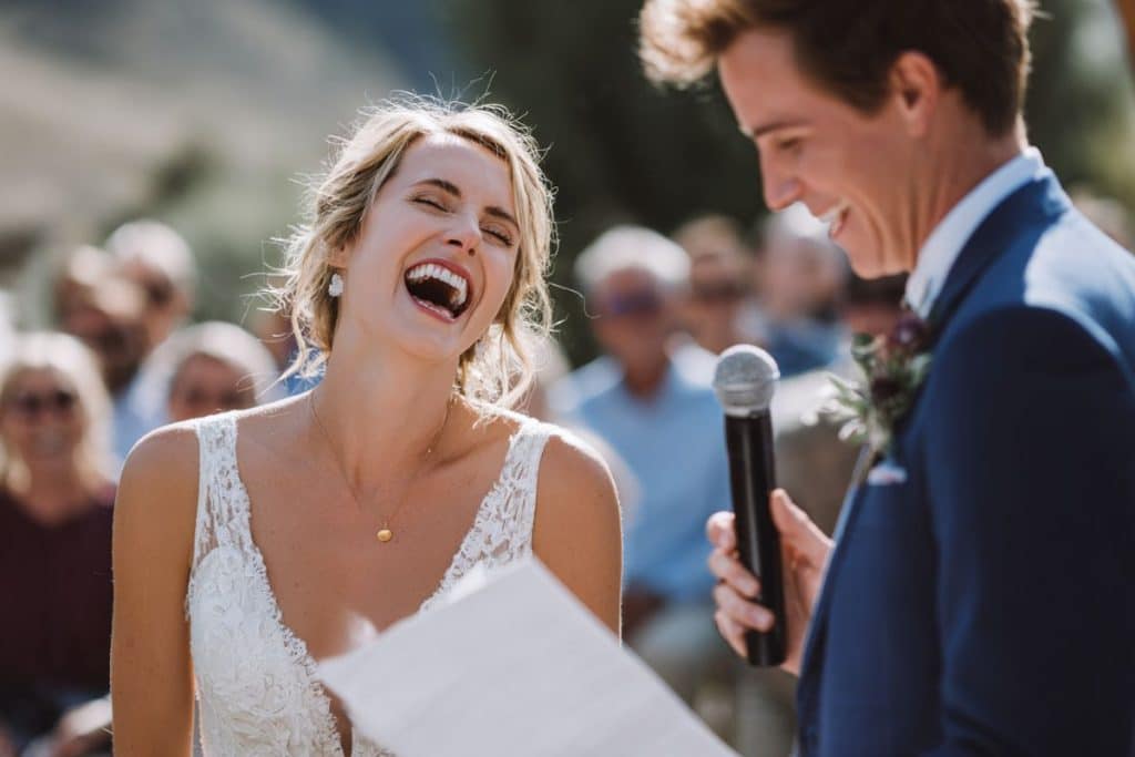 Mariée éclatant de rire pendant le discours humoristique du marié lors d'une cérémonie laïque en extérieur