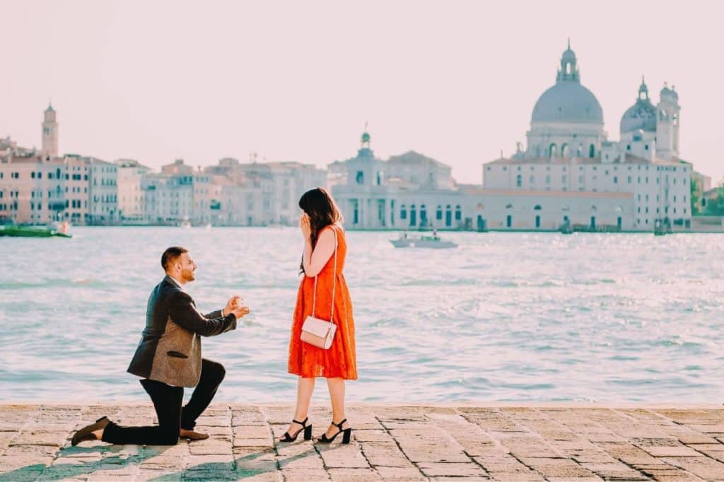 homme à genoux pour demander en mariage sa fiancée robe rouge venise italie bord de l'eau