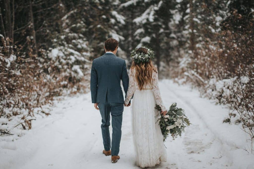 Couple de mariés marchant main dans la main dans une allée enneigée bordée d'arbres vers un château français au coucher du soleil hivernal