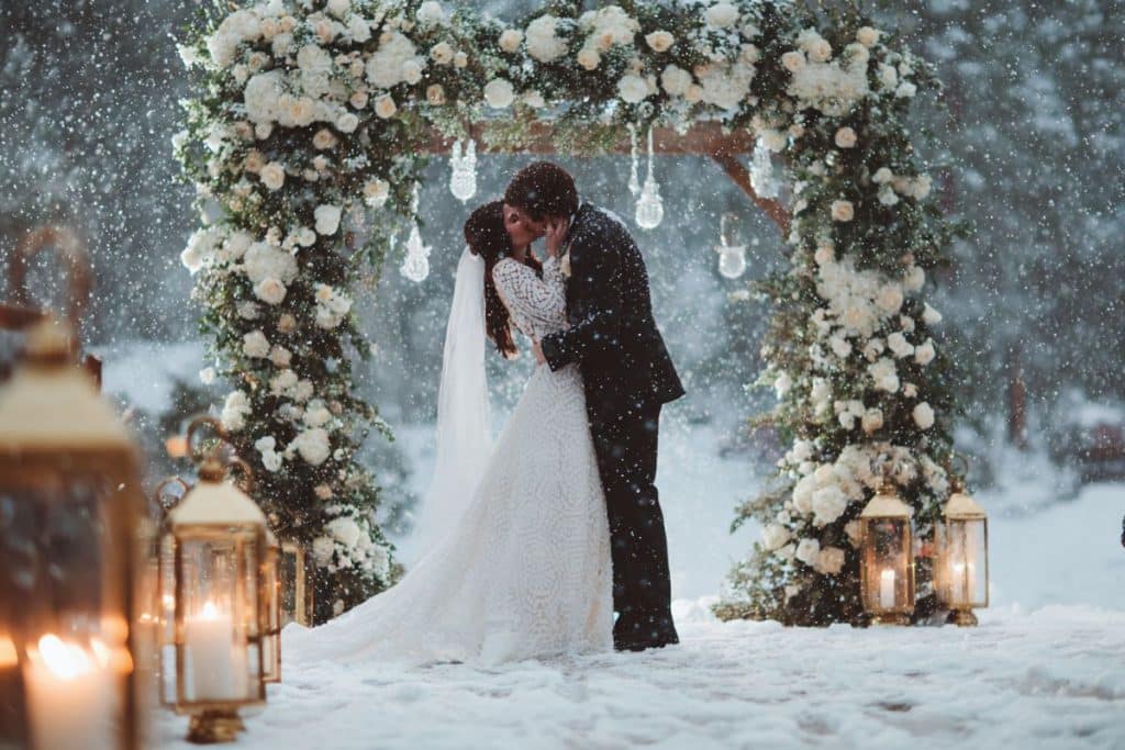 Premier baiser de mariés sous la neige avec arche fleurie de roses blanches et lanternes vintage dorées au coucher du soleil