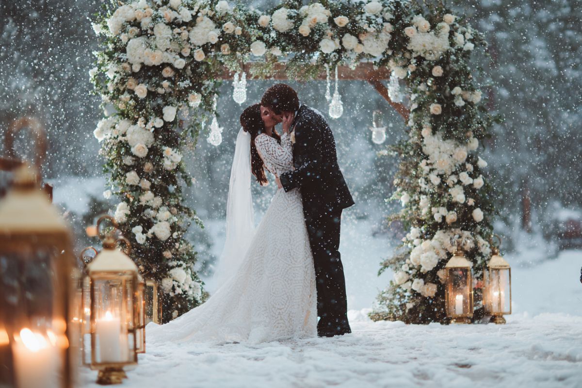 Premier baiser de mariés sous la neige avec arche fleurie de roses blanches et lanternes vintage dorées au coucher du soleil