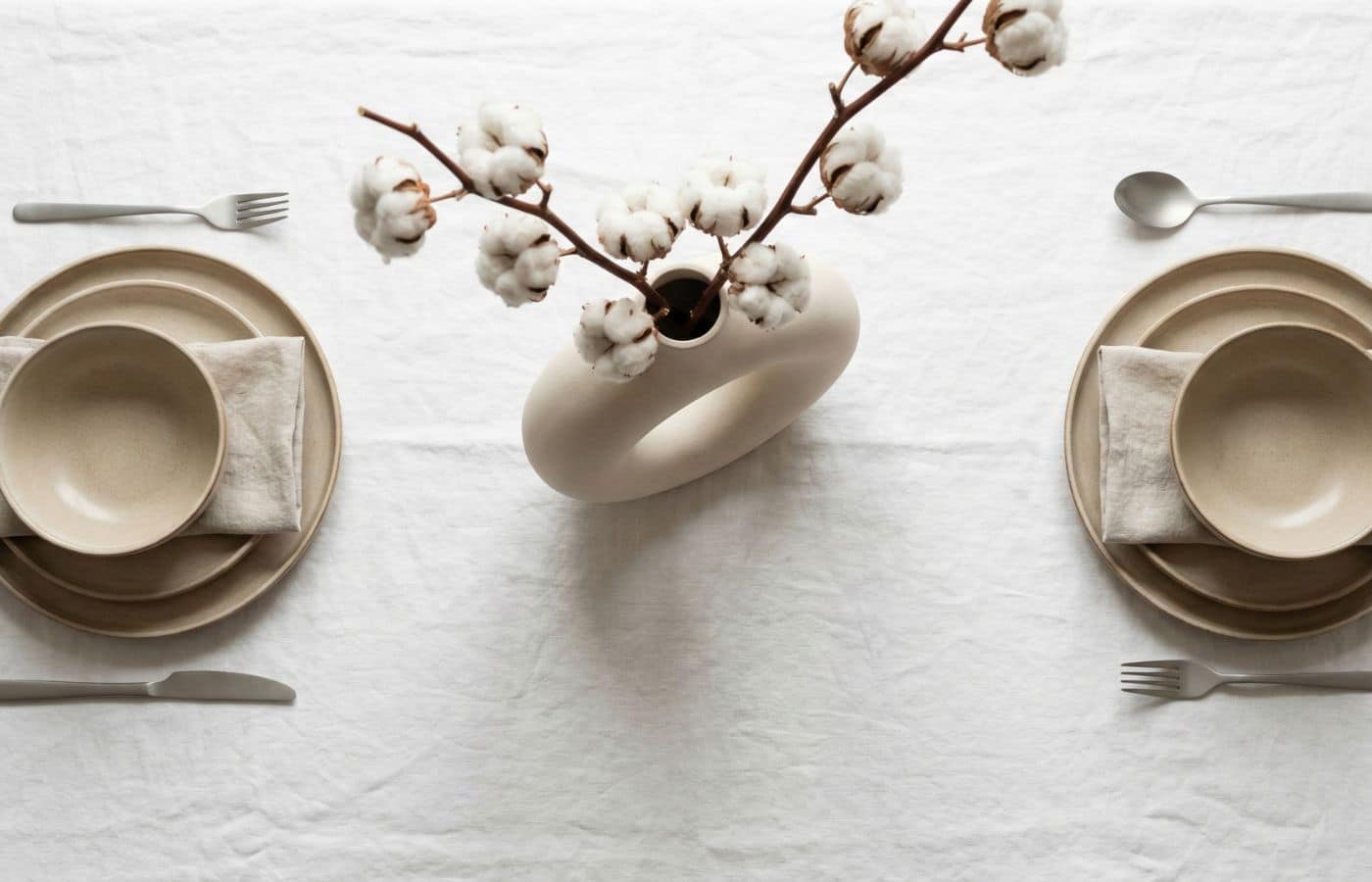 Vue de dessus d'une décoration de table de mariage bohème minimaliste avec nappe en lin blanc, vaisselle en grès beige et branche de coton.