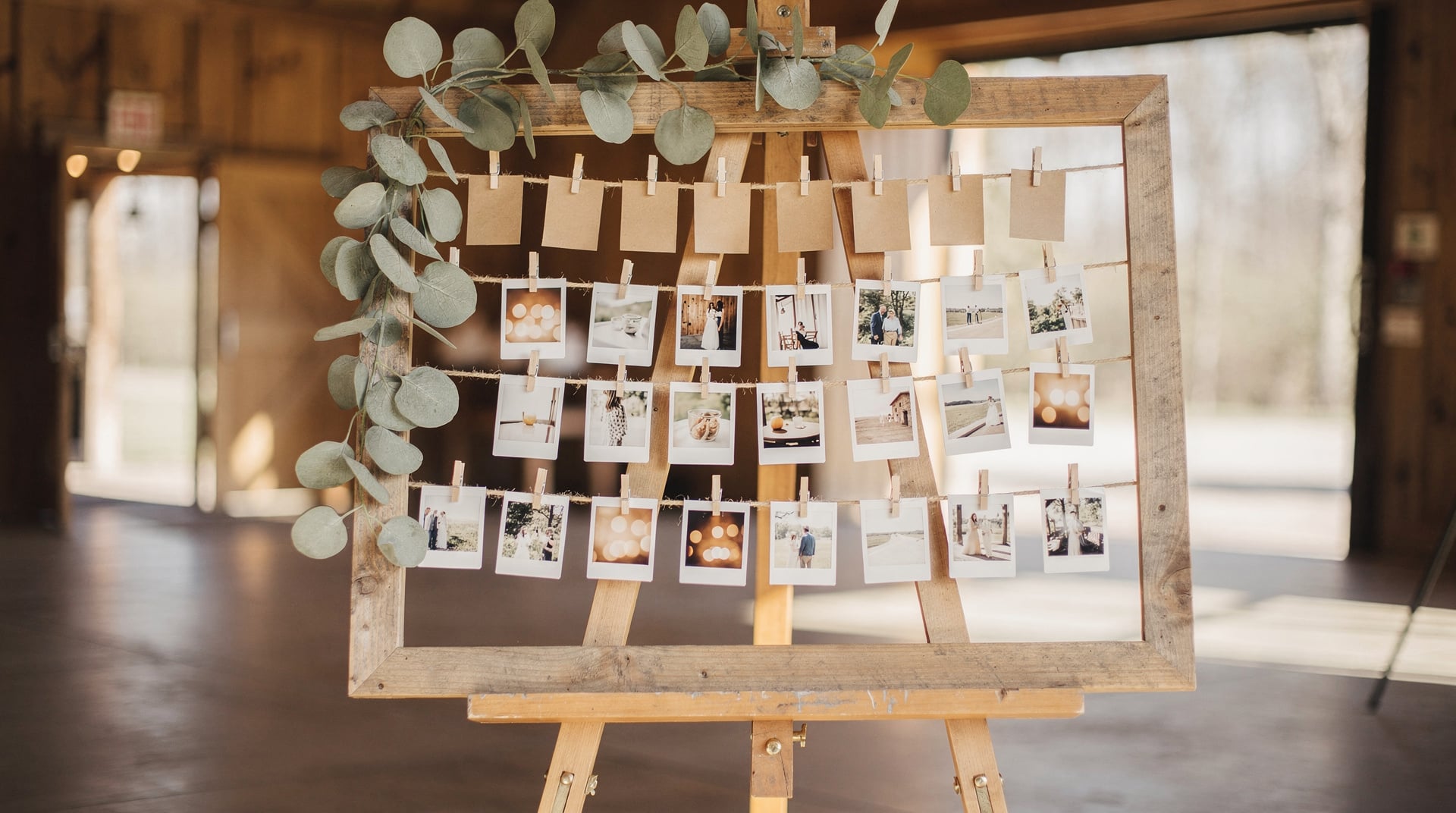 Plan de table mariage en photos : cadre en bois sur chevalet, trois rangées de tirages instantanés pincés sur de la ficelle sous des étiquettes kraft, guirlande d'eucalyptus sur le coin du cadre