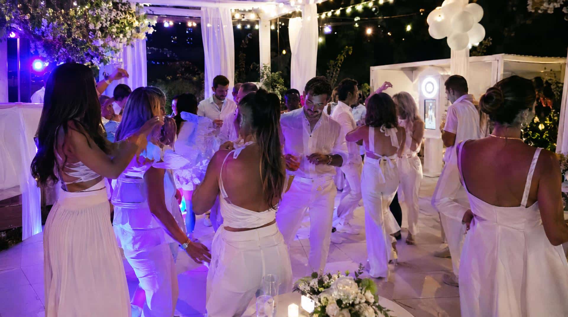 Invités en blanc dansant sous la lumière noire lors d'une soirée blanche, photobooth blanc en fond